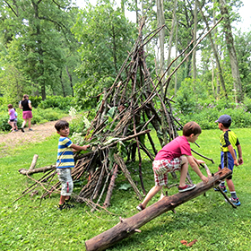 Boys making stick shelter while other children explore a path in the woods.